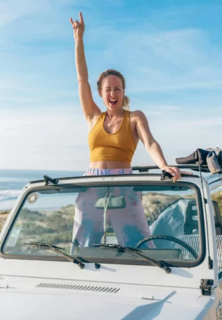 Joyful woman in a yellow tank top standing through the sunroof of a white car, raising her hand in a peace sign, with a beach in the background.